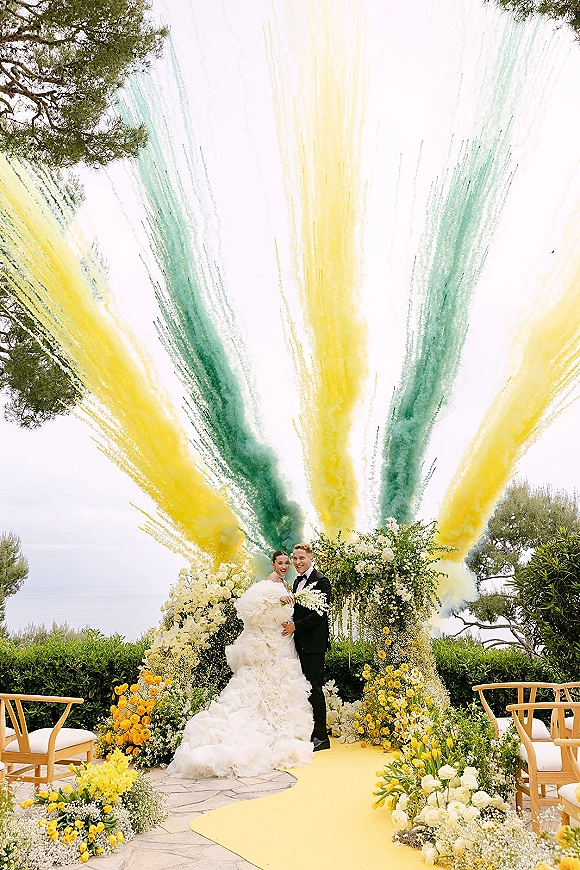 Ceremony moment with wedding smoke bombs as bride and groom stand by a floral arch on a yellow aisle runner overlooking the ocean view