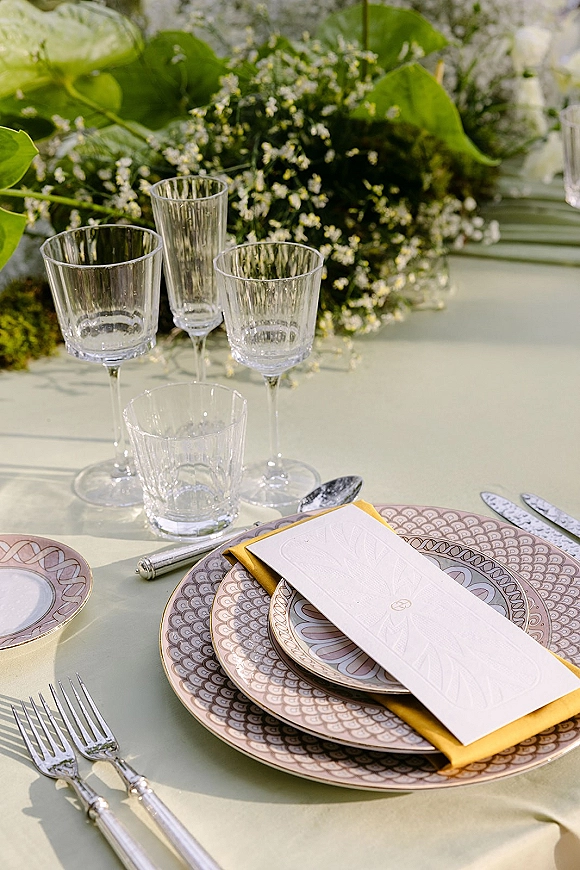 Reception tablescape with wedding place setting, lilac patterned plates, yellow napkin, menu card, clear goblets, and floral centerpiece on pale green linen outdoors