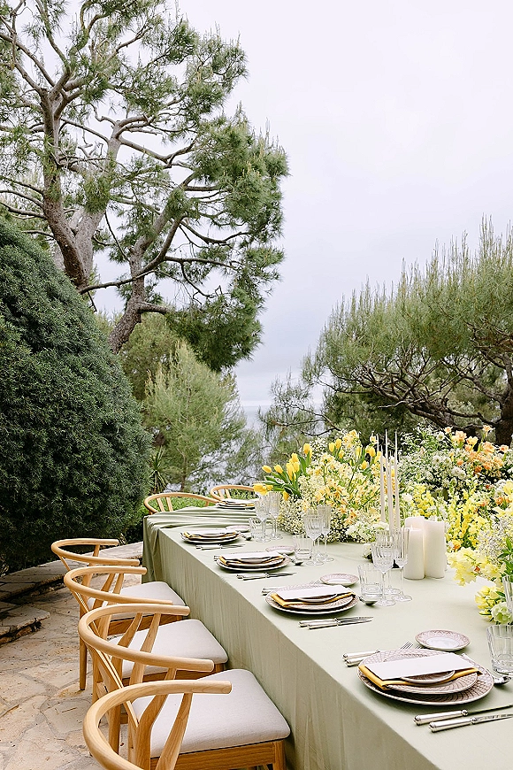 Reception tablescape with an outdoor reception table on a stone patio, sage green tablecloth, yellow and white florals, candles, crystal glassware, and ocean view