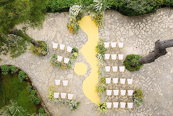 Ceremony aisle design with a yellow aisle runner curving through white chairs, lined with white and yellow florals on a stone patio garden setting