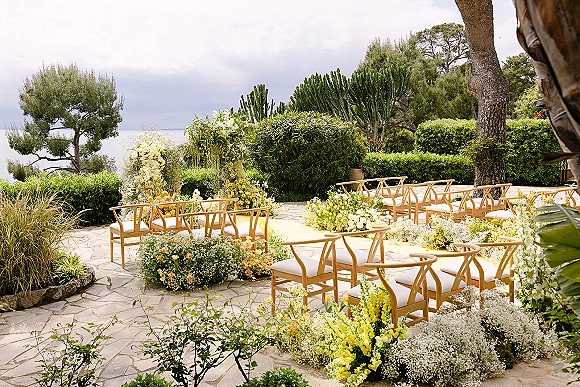 Ceremony setup with wood chairs and a flower-lined aisle runner leading to a yellow-and-white floral arch on a stone patio with ocean view