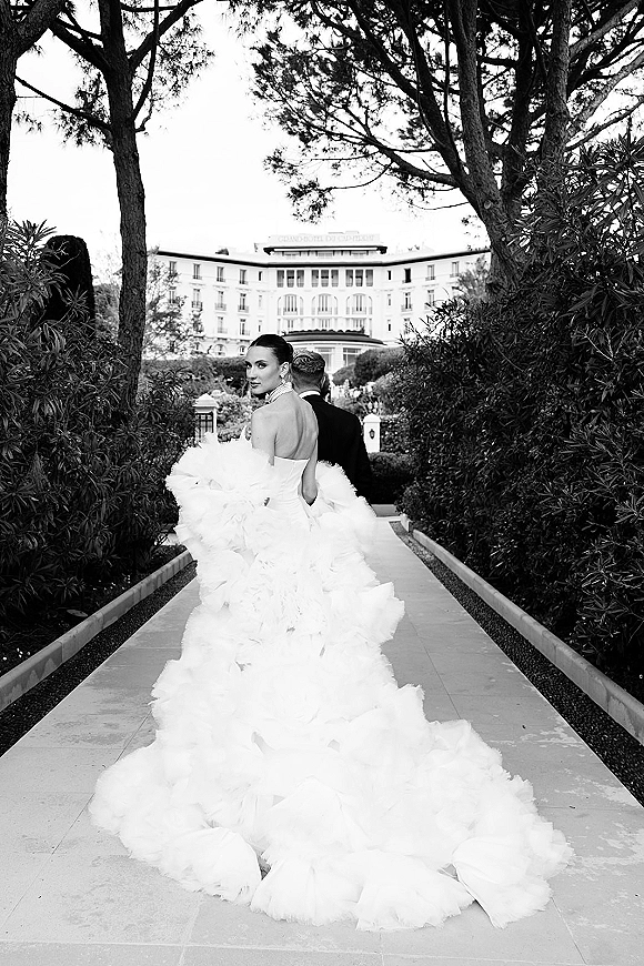 Couple portrait in a black and white wedding portrait, bride looking back as they walk a garden path past a grand building facade
