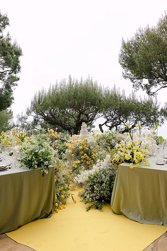 Reception tablescape with an outdoor reception table set in olive green linens, yellow and white floral centerpieces, candles, flutes, and cake on patio under trees