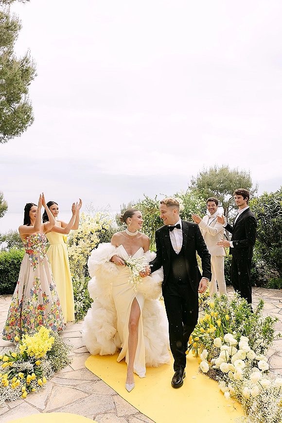 Wedding recessional as bride and groom walking aisle under a floral arch, bride holding white bouquet, guests clapping on garden path
