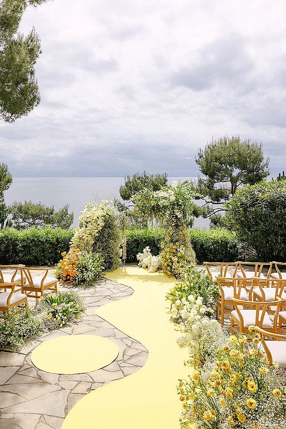 Ceremony aisle design with a yellow aisle runner leading to an asymmetrical floral arch on a stone patio overlooking the ocean under cloudy skies