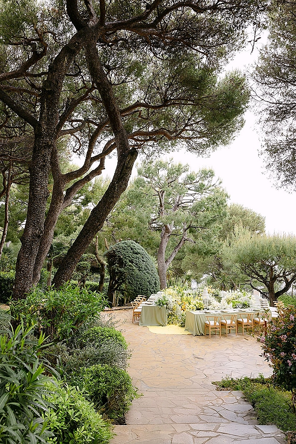 Outdoor reception setup with a long banquet table in sage green linens, yellow and white florals, and candles on a stone patio with pine trees