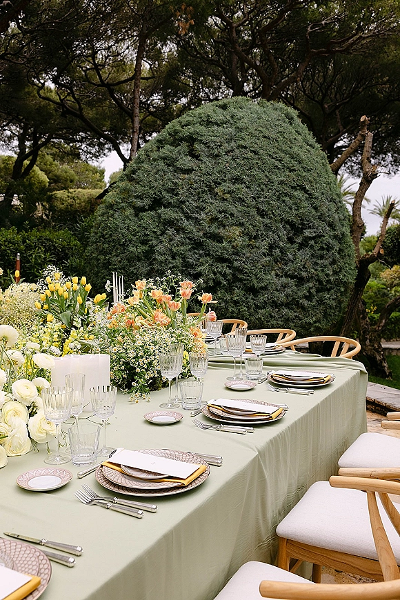 Reception tablescape with sage green tablecloth, tulip and greenery garland centerpiece, taper candles and patterned place settings on a garden lawn