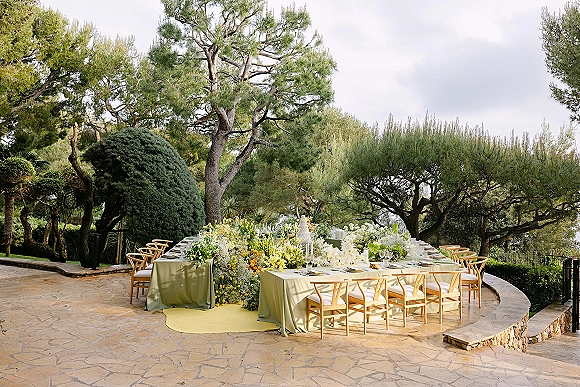Reception tablescape with a long outdoor reception table in green linens, yellow and white florals, candles, and place settings on a stone patio