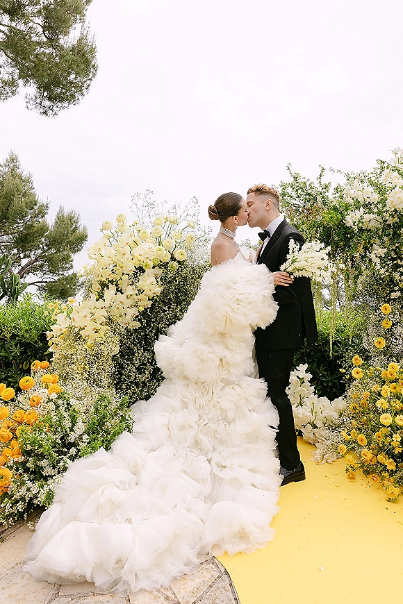 Wedding kiss portrait of bride and groom kissing beneath a floral arch, her strapless ruffled gown with long train flowing in a garden setting