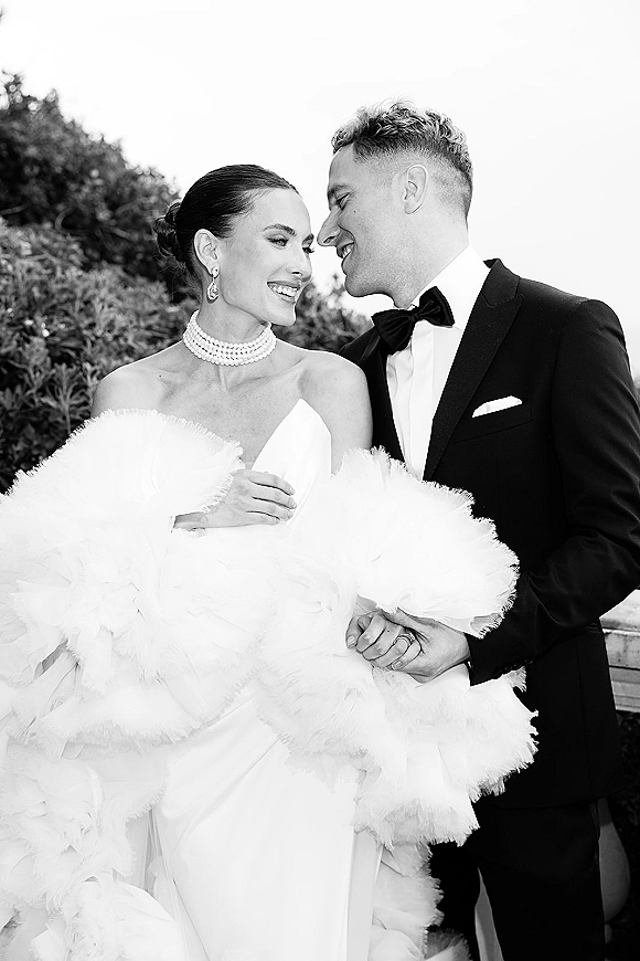 Couple portrait in a black and white wedding portrait, bride in strapless gown with pearl choker holding hands with groom in tux by stone railing