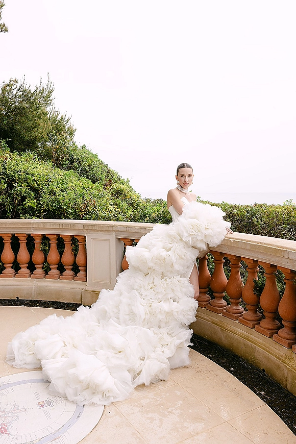 Bridal portrait of a bride in a ruffle wedding dress with a dramatic train, leaning on a stone terrace railing with ocean view behind her