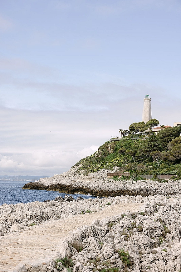 Coastal lighthouse view with a white lighthouse on a cliff and stone path, overlooking rocky coastline and ocean under cloudy sky