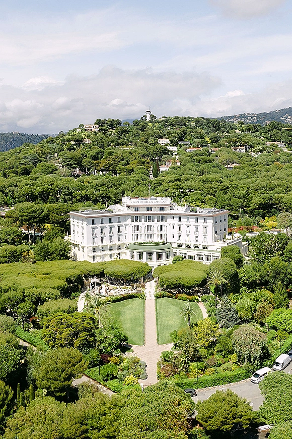 Wedding venue exterior with a luxury wedding venue grand hotel, manicured lawn and palm-lined pathways, set against hillside forest and clouds