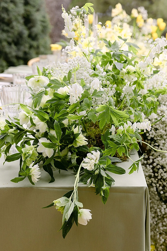 Wedding centerpiece of white flowers and greenery with baby’s breath, trailing foliage, and glass votives on a linen table in a garden setting
