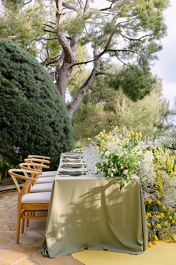 Reception tablescape with a green tablecloth on a long banquet table, yellow tulip centerpiece, gold flatware, set on a stone patio