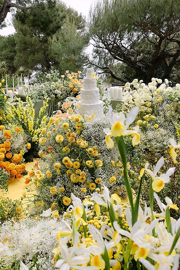 Wedding cake display with a tiered white wedding cake on green linens, surrounded by yellow and white blooms and taper candles in a garden setting