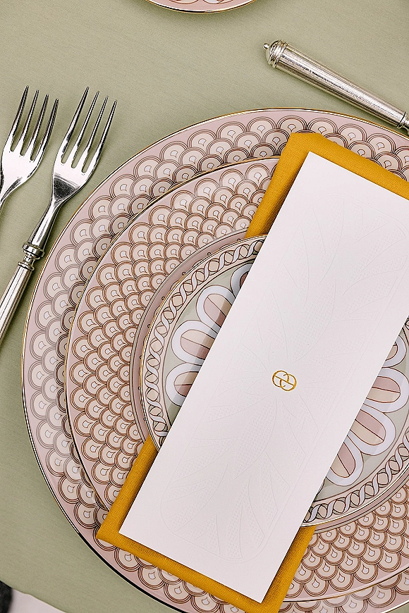 Wedding place setting with wedding tablescape details, patterned and gold-rim plates, mustard napkin, menu card, and silver flatware on sage green tablecloth
