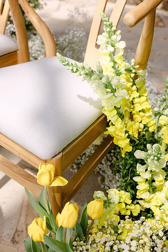 Ceremony chair florals with yellow and white tulips and snapdragons beside a wood chair on a stone floor in outdoor light