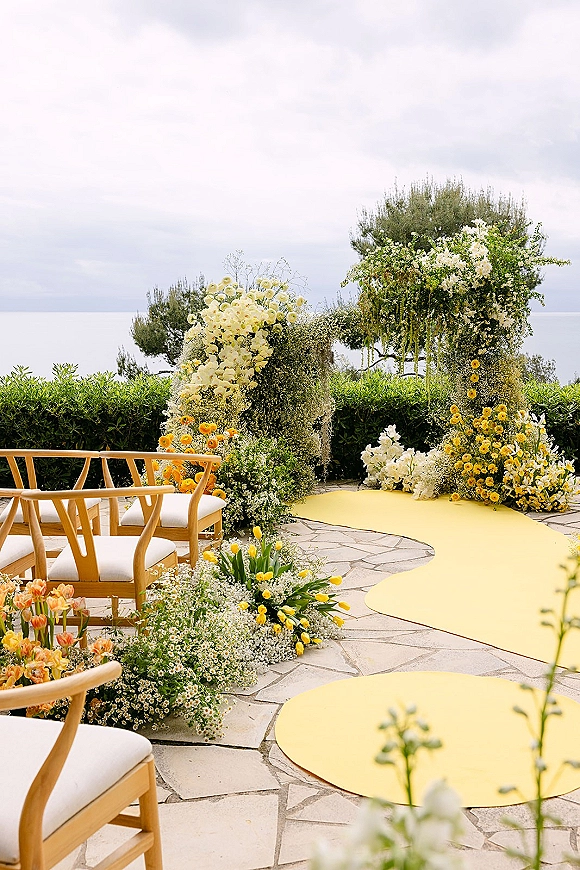 Ceremony setup for an outdoor wedding ceremony with a floral arch, yellow aisle runner, and wooden chairs on a stone patio with ocean view