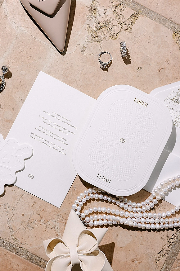 Wedding invitation suite with embossed wedding invitations in gold foil, styled with engagement ring and pearls on sunlit stone surface