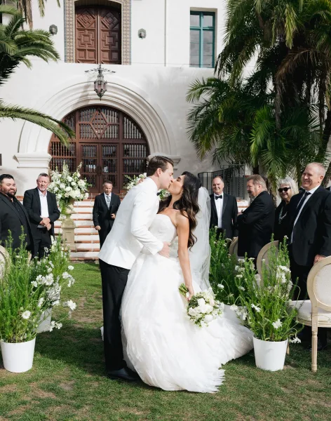 Wedding kiss moment as the couple shares a ceremony exit kiss beneath a white stucco arched doorway with palm trees, her veil trailing.