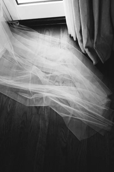 Wedding veil detail of a tulle wedding veil draped on a hardwood floor by a window with soft curtains, highlighting its sheer fabric.
