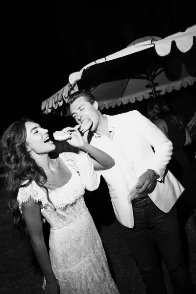 Reception moment with a couple sharing dessert as the bride feeds the groom an ice cream sandwich under a night sky on the outdoor lawn.