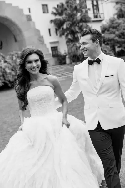 Couple portrait of the bride and groom walking, smiling and holding her skirt, his black bow tie neat, past palm trees and an archway.