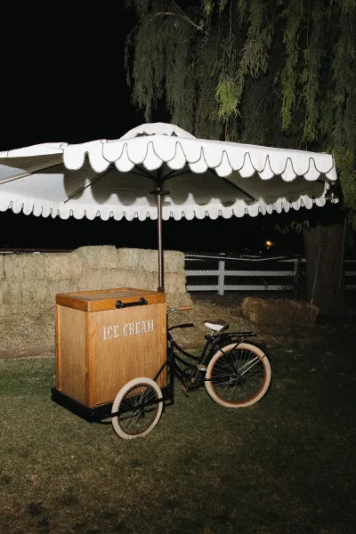 Ice cream cart serving as a cozy wedding dessert station, shaded by an umbrella on a grass lawn with hay bales and a white fence at night.