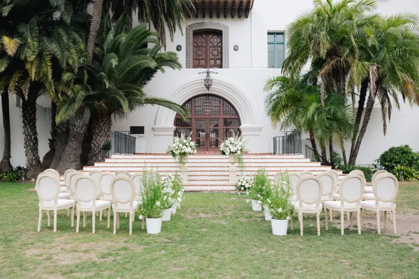 Ceremony setup with outdoor ceremony seating and white florals facing arched wooden doors on brick steps, framed by palm trees and stucco facade.