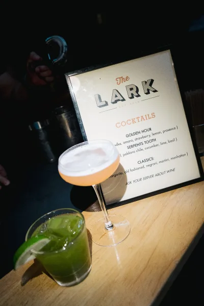 Cocktail menu sign as a wedding bar sign beside a coupe glass on a wood bar top, with a dark bar backdrop and a cocktail shaker and strainer.