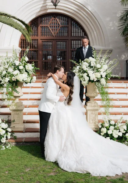 Wedding first kiss as the bride and groom kiss on stone steps, framed by arched wooden doors, her long veil trailing.
