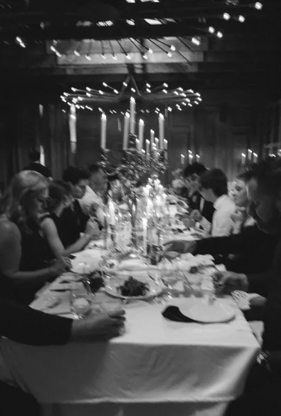 Reception dinner moment with wedding guests dining at a long table, taper candles glowing beneath wood ceiling beams and draped walls.