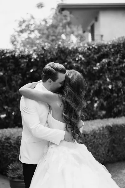 Couple portrait as a bride and groom hug by a garden hedge and shrubs, her bridal gown brushing the path near the house exterior.