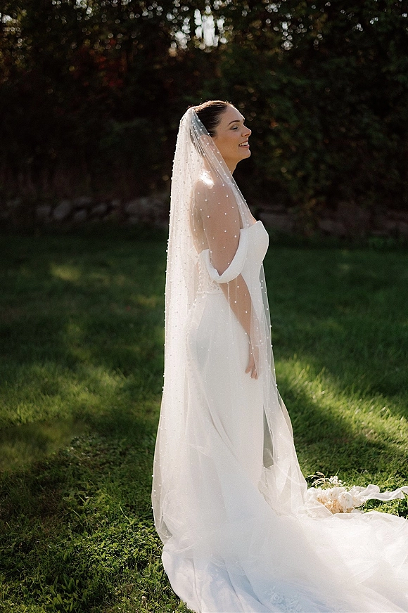 Bridal portrait of a side profile bride smiling in an off-the-shoulder wedding dress, pearl cathedral veil glowing in sunlit trees