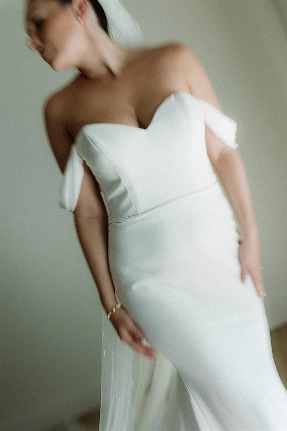 Wedding dress close-up of an off the shoulder wedding dress with strapless neckline, veil, earrings, and bracelet against a neutral wall
