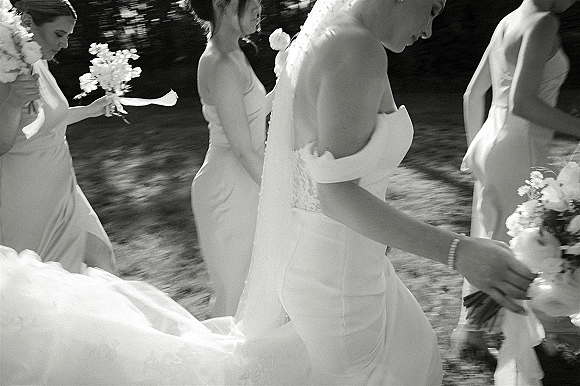Bridesmaids walking together in a candid moment, holding bouquets beside the bride’s long veil on a sunlit lawn with trees