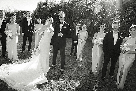 Wedding party portrait with bride and groom centered, bridesmaids holding white bouquets and groomsmen in black suits on a sunlit lawn with flare