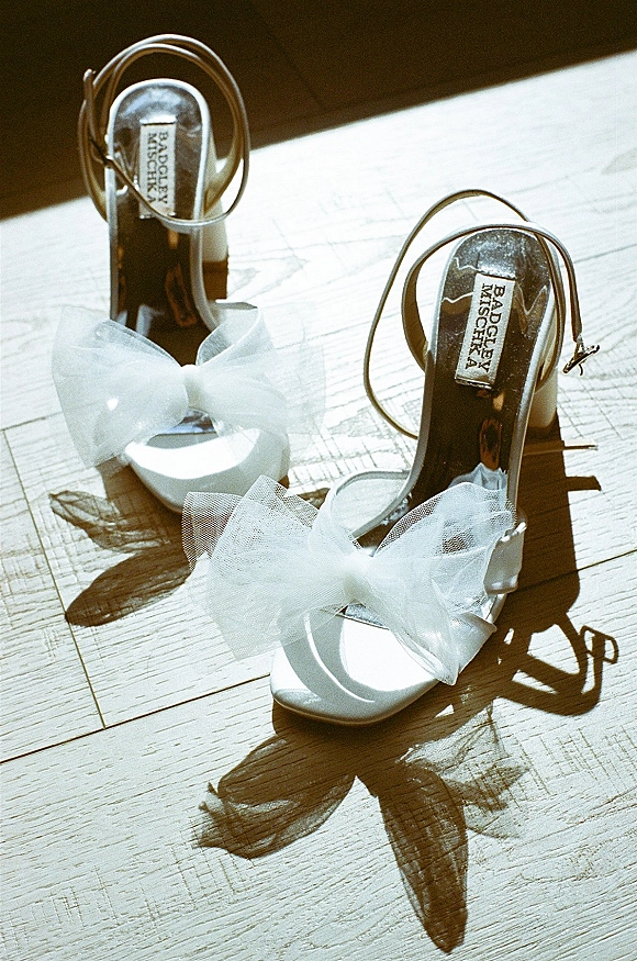 Wedding shoes in white high heel sandals with tulle bows and ankle straps, metallic insoles catching sunlight on a wood floor