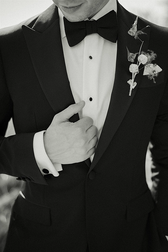 Groom portrait in a black tuxedo groom look with bow tie and boutonniere, dress shirt and cufflinks against a blurred outdoor background