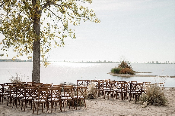 Ceremony setup with pampas grass arrangements beside rows of wood crossback chairs on a sandy lakefront under a large tree