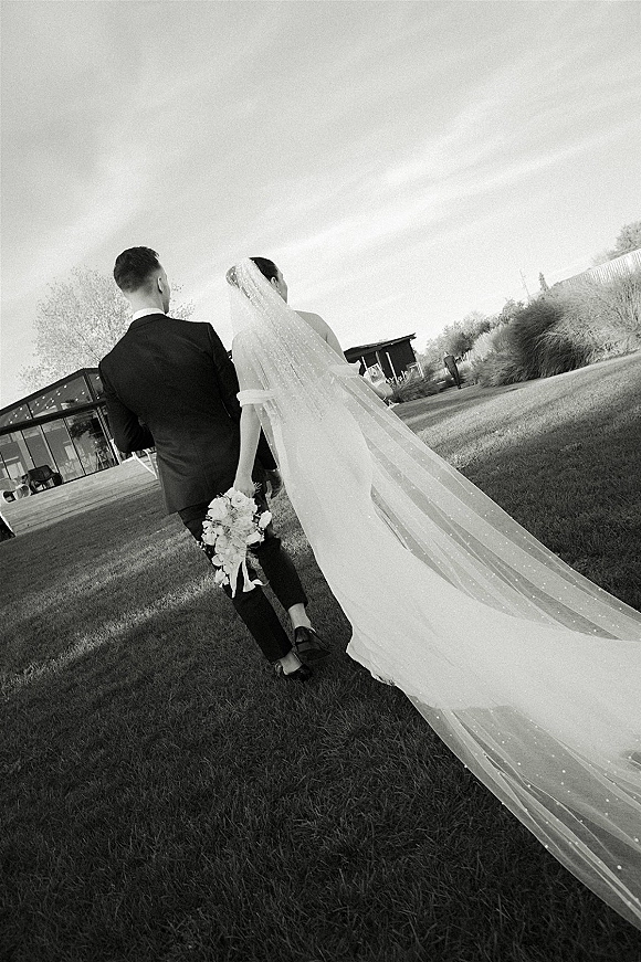 Couple portrait in a black and white wedding photo, bride and groom walking away on a lawn by a modern building, her long veil trailing