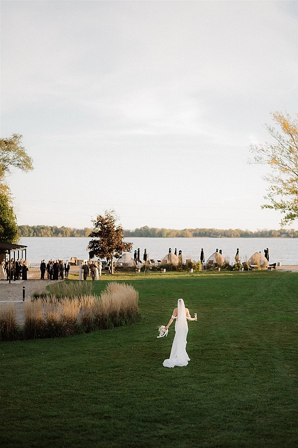 Bridal portrait of a bride walking away on a lakeside lawn, long veil flowing, holding a white bouquet and champagne flute near mingling guests
