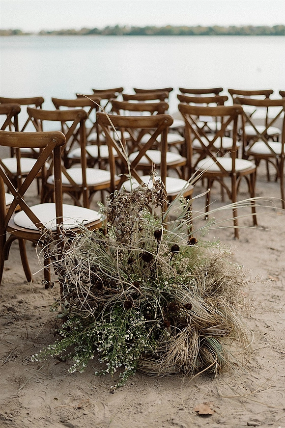 Ceremony seating with wood cross-back chairs and white cushions in rows on sandy beach by lake, with dried grass aisle flowers