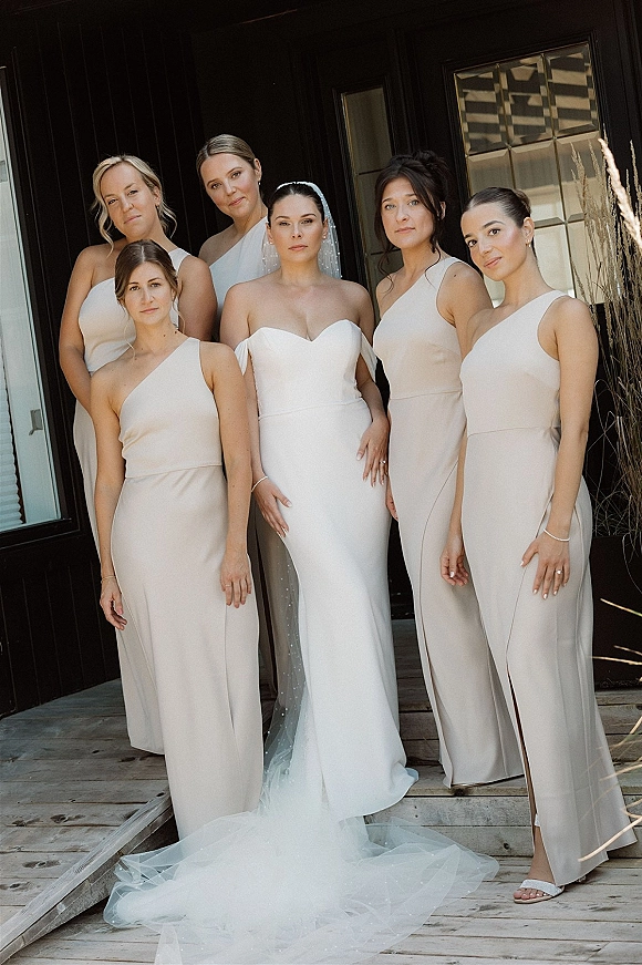 Bride and bridesmaids in neutral dresses pose in front of a black wall, the bride in a strapless gown with a long veil train
