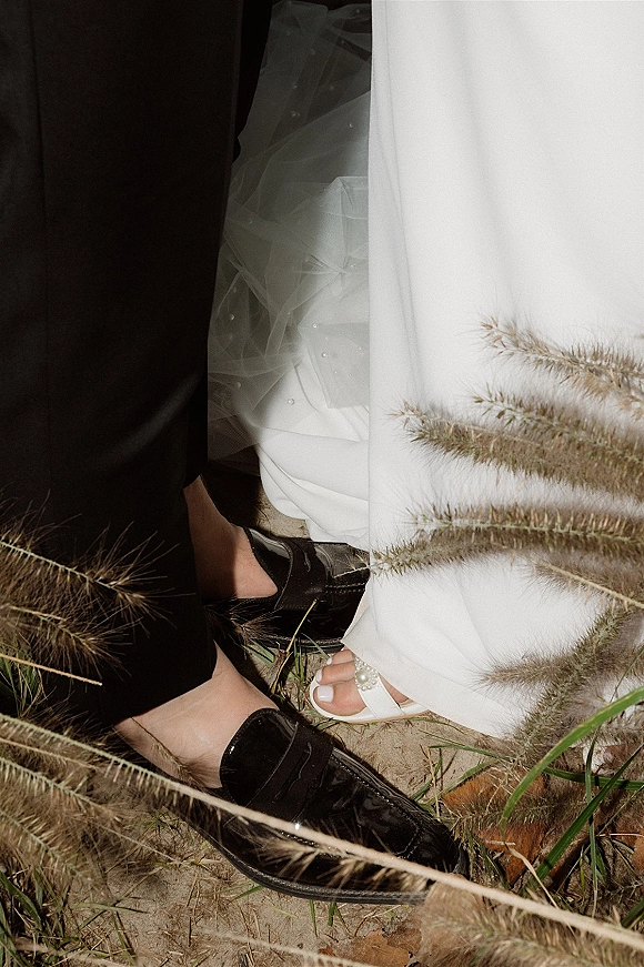 Wedding shoe detail showing white bridal heels with pearl accents beside black groom loafers, tulle veil and dress hem on sand with pampas grass