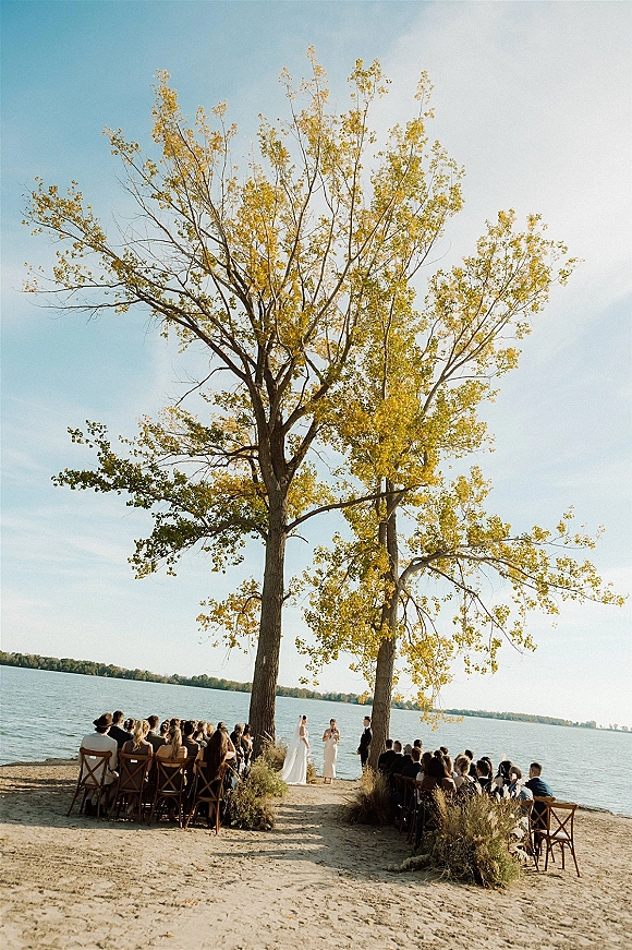 Outdoor wedding ceremony with lakeside wedding ceremony aisle greenery, wooden chairs facing a floral arch on sandy shore under tall trees