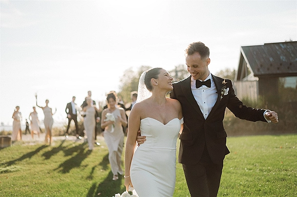 Wedding recessional as bride and groom walk hand in hand across a sunlit lawn toward a rustic barn, her veil flowing behind him in tuxedo