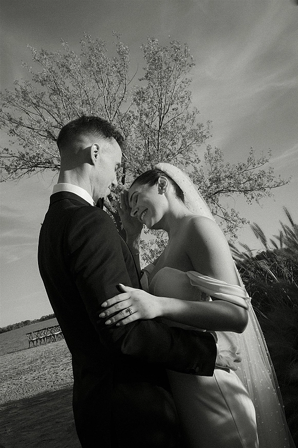 Couple portrait, black and white wedding portrait of bride and groom embracing with forehead touch by a pier beside calm water and trees