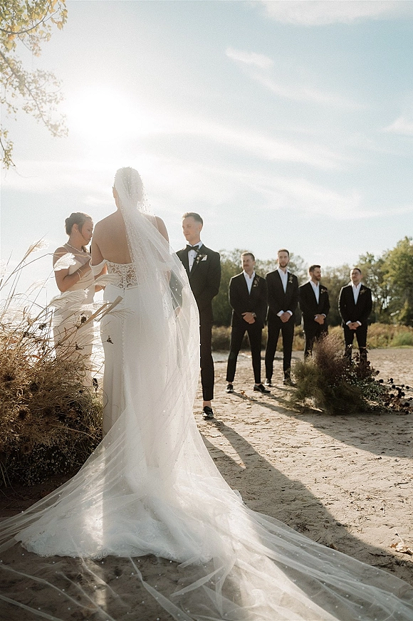 Ceremony moment as the bride walks to groom during an outdoor wedding ceremony, cathedral veil flowing on a sunlit path in tall grass